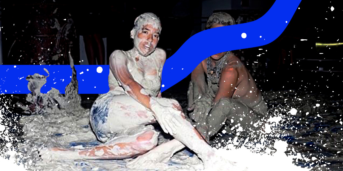 A short-haired dyke sits on a mud spattered floor with one leg folded under them and the other extended. They smile an easy grin into the camera and place a hand on their extended knee. Behind them a blue graphic ribbon runs behind them and across another mud covered person sitting behind them.