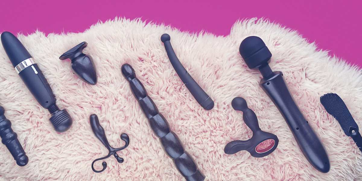 A range of matte black sex toys, from a wand vibrator to a butt plug to a curved g-spot wand to a light crop-style spanking paddle, are arranged artfully on a lush faux fur rug against a hot pink background.