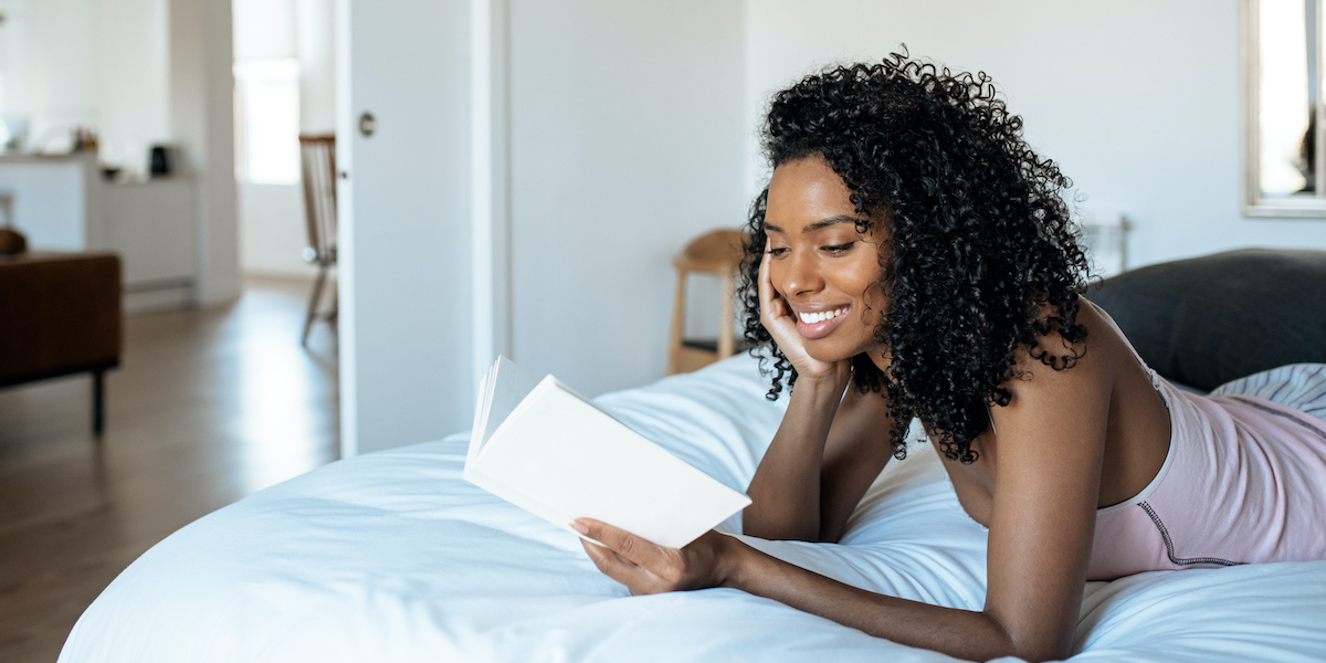 a Black woman reading a book on top of the covers of a bed