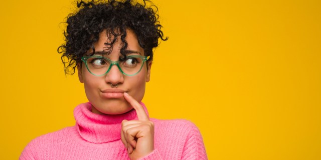 A Black person with loosely curled hair wearing quirky green-framed glasses, a septum piercing and a bright pink turtleneck looks off to the side nervously while tapping their mouth in a thinking gesture