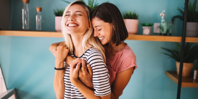 One thin white woman hugs another thin white woman from behind while both grin with their eyes closed, besotted, and clutch each others' hands in a well-lit and attractively decorated apartment.