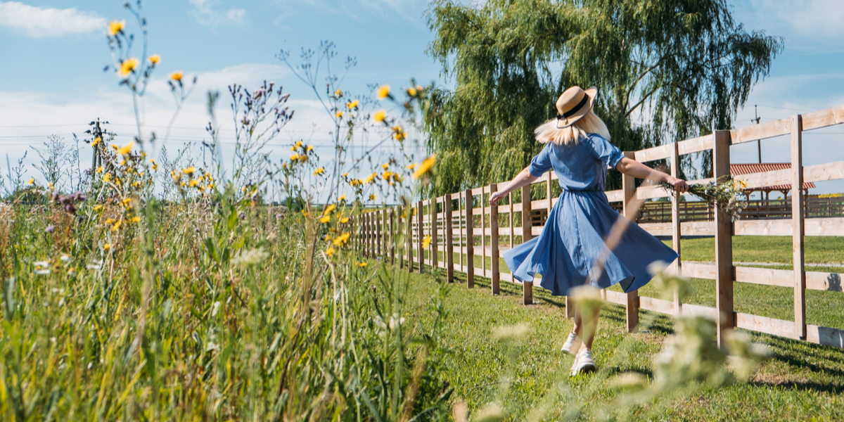 A woman in a blue dress and straw hat with blond hair walks alongside a fence in a grassy field.