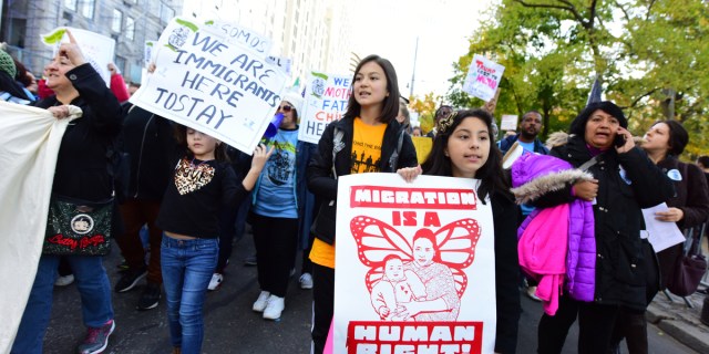 A pro-migrant protest or rally, with multiple people marching and holding signs; two visible ones read "Migration is a human right" and "we are immigrants here to stay."