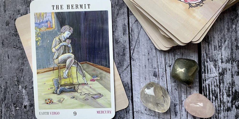 A stack of tarot cards on a weatherworn wooden table with the Hermit card facing up and three small crystals beside the deck
