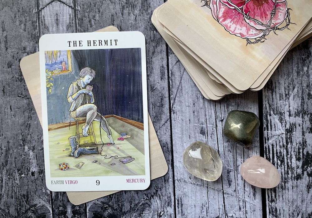 A stack of tarot cards on a weatherworn wooden table with the Hermit card facing up and three small crystals beside the deck