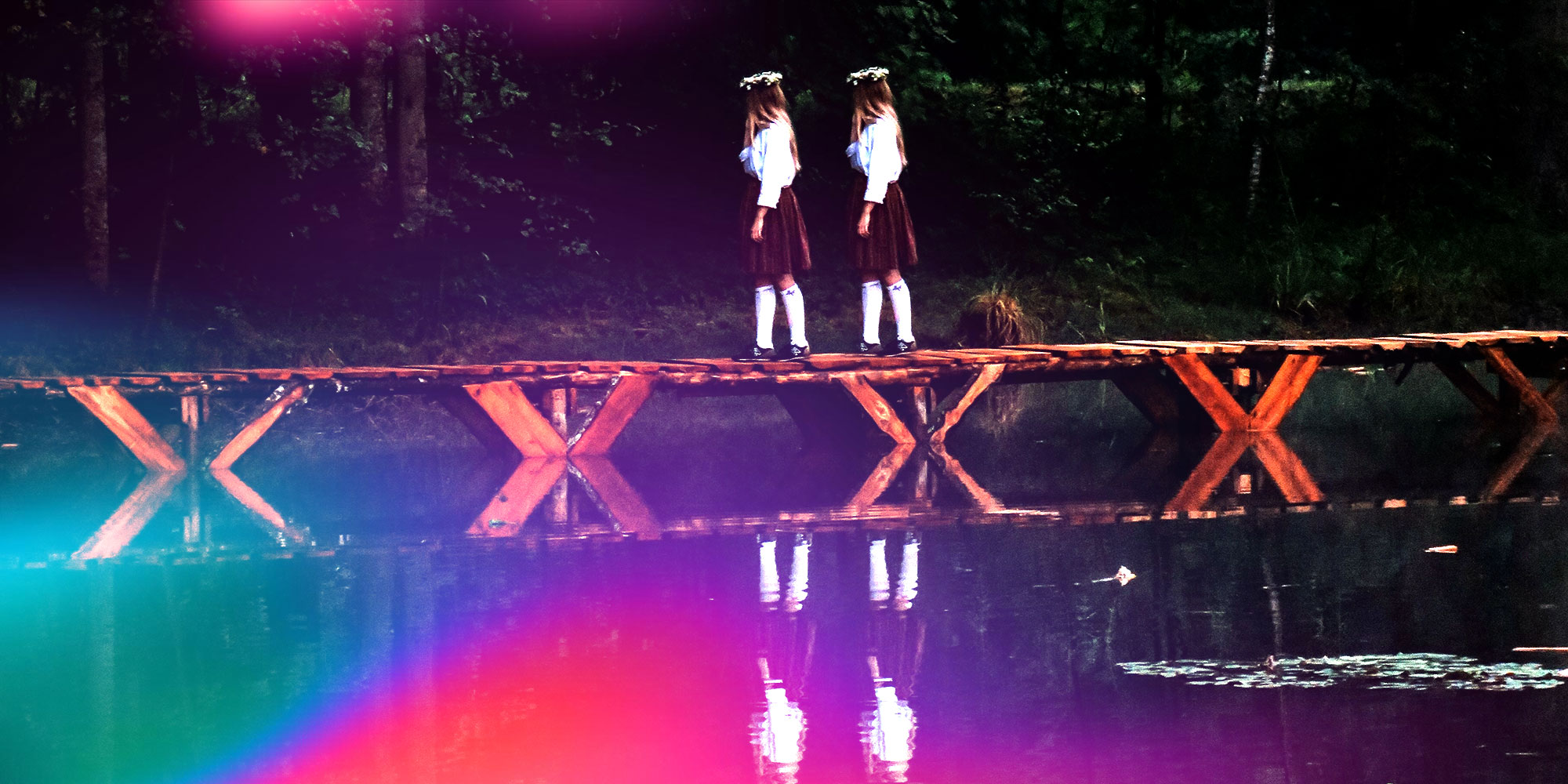 two girls in school uniforms walking across a bridge over the water in a forest