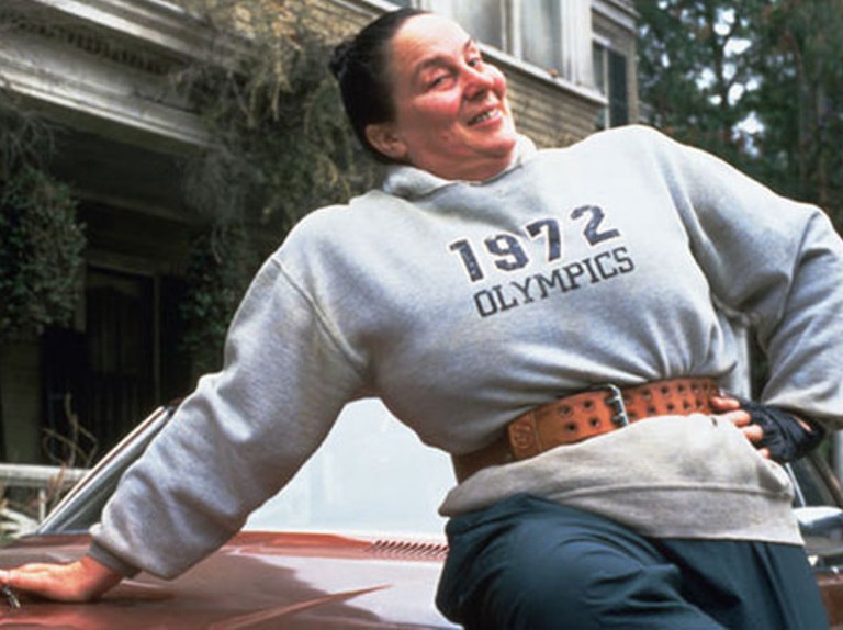 Miss Trunchbull from the Matilda movies leans back across the hood of a car, wearing a gray sweatshirt that reads 1972 OLYMPICS, a weightlifting belt and gloves