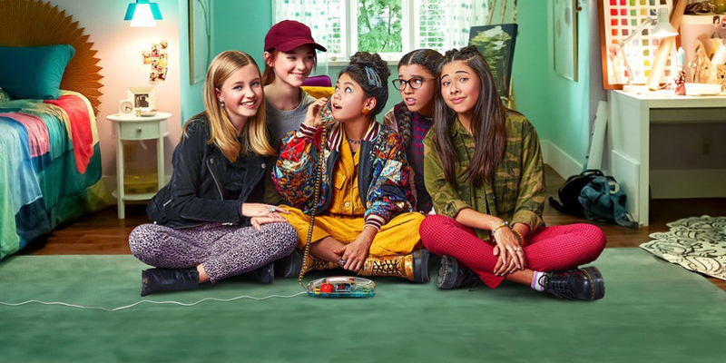 The cast of Netflix's The Baby-Sitters Club sit together on the floor a green bedroom. Claudia is in the center of the group, and she talks on a clear phone from the 1990s.
