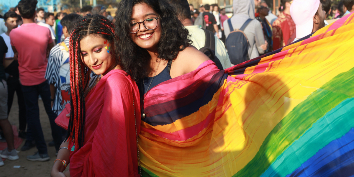 Two South Asian queer femmes with long hair embrace underneath a rainbow flag.