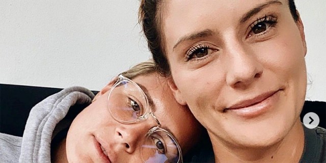 Ali Krieger and Ashlyn Harris smile directly into the camera. Ashlyn is wearing a grey hoodie and clear glasses, she rests her head on Ali's shoulder.