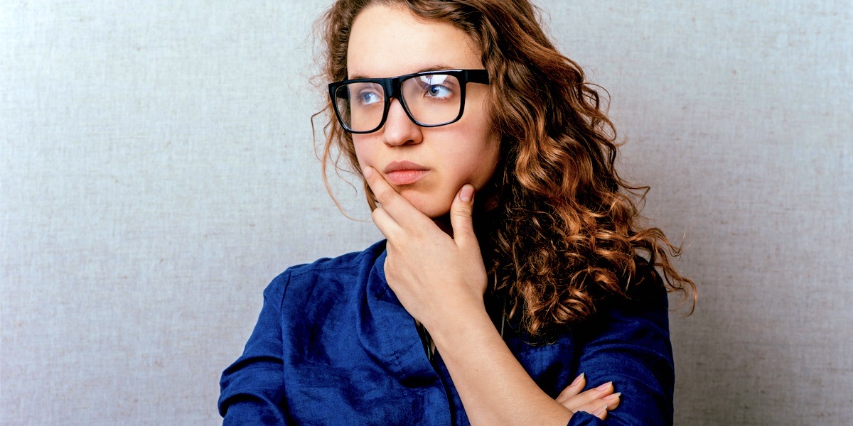 A person with long curly red hair and a very cute pair of thick black glasses, wearing a dark blue denim shirt, stares off to the left of the camera and thinks hard about their ex.
