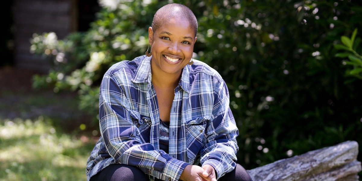 A photo of Angela Walker, a Black woman with a shaved head, seated on a log, leaning forward and smiling broadly in a blue checked flannel shirt