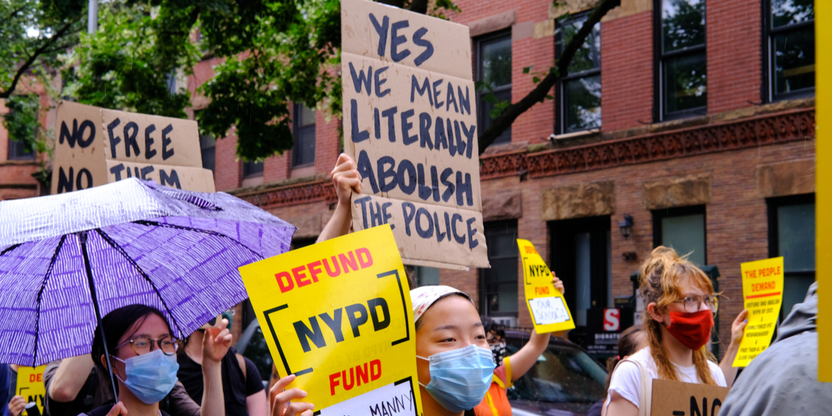a group of protestors in masks, one of them holds a sign that says "yes we literally mean abolish the police" and another says "defund NYPD"