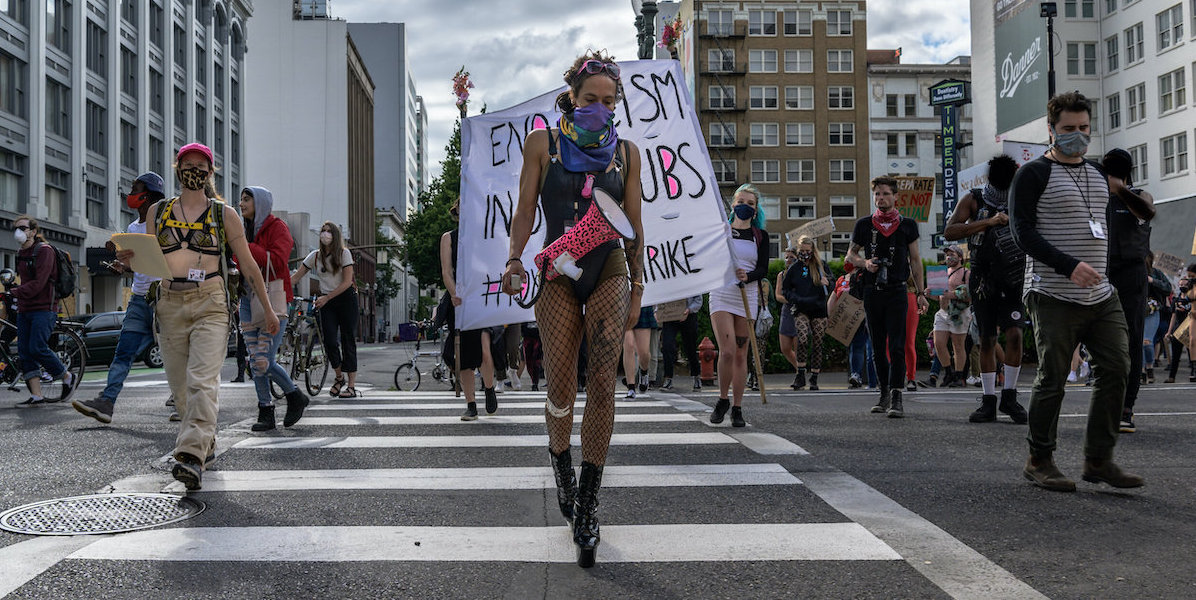 a group of people protesting in the street and soberly walking across a crosswalk. the person in the center wears black boots, black fishenets, a black body suit, a lilac scarf over their face and is carrying a hot pink and black cheetah megaphone across their body. they look downcast. behind them a protest sign half blocked by the people walking says "end racism in strip clubs #pdxstripperstrike"