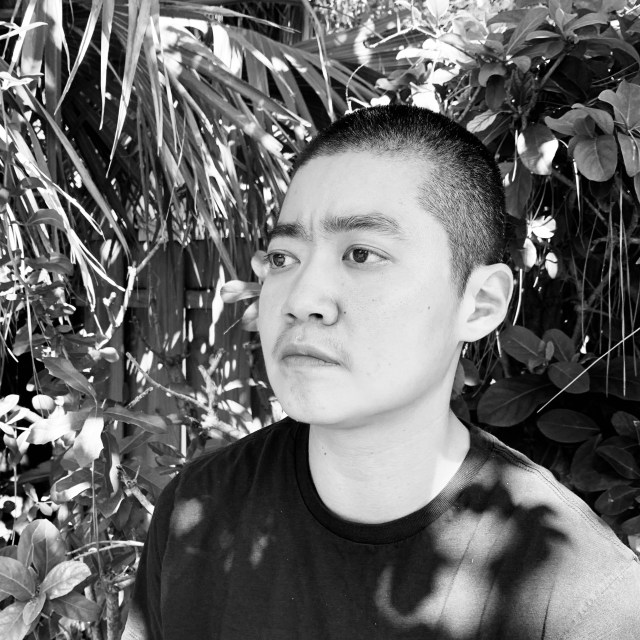 black and white photo of patty yumi cottrell, a trans korean adoptee, looking somberly into the distance. they have close-cropped hair and are wearing a black t-shirt as they stand in front of a thicket of palms leaves.