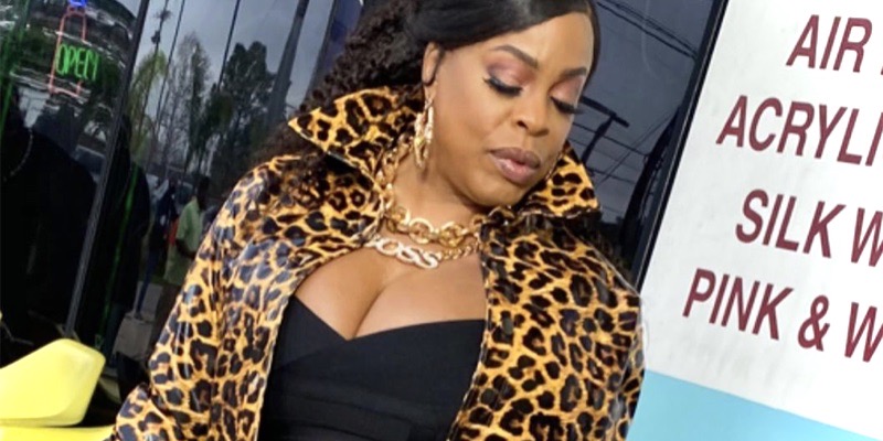 Niecy Nash stands against a black window in a low cut black body suit and a leopard print jacket. Her hair is pulled back into a low ponytail.