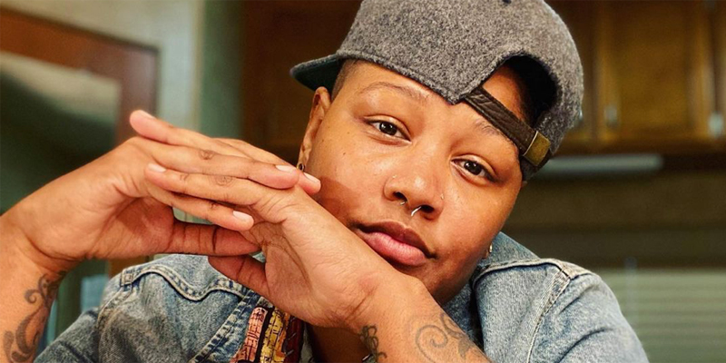 Rahne Jones has a backwards cap and sits in a chair with her hands clasped in front of her face and her tattoos showing.