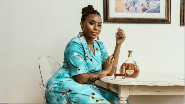 Chef Kia Damon, a black woman in a turquoise short sleeve dress and dreadlocks piled back into a top knot bun, sits at a wood table next to a bottle of whiskey.