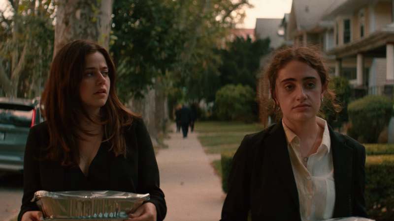 Rachel Sennott and Molly Gordon walk next to each other outside. Molly is carrying a tin food tray and looking at Rachel whose makeup is smeared.