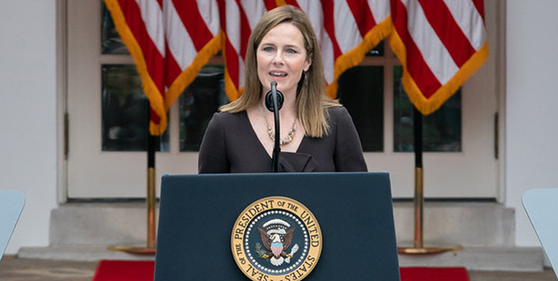 amy coney barrett, a white woman with shoulder length brown hair, stands in front of a podium, with a back drop of american flags behind her. her mouth is open and her eyes are squinting as she speaks.