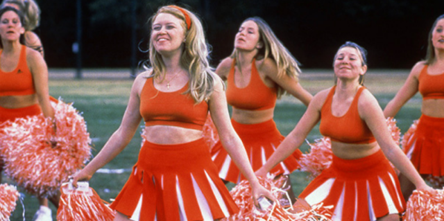 A group of cheerleaders in red-orange uniforms with pom poms cheer.