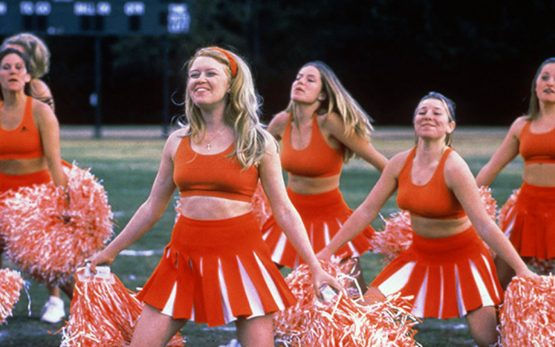 A group of cheerleaders in red-orange uniforms with pom poms cheer.