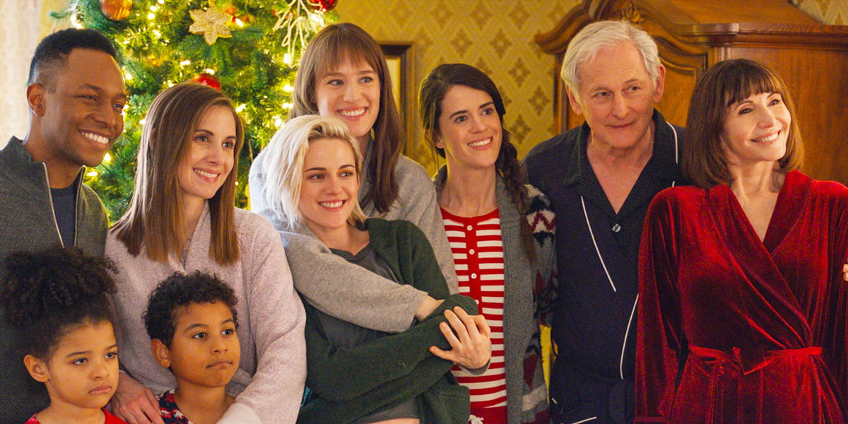 A still from Happiest Season with Mackenzie Davis' arm around Kristen Stewart with her family in front of the Christmas tree