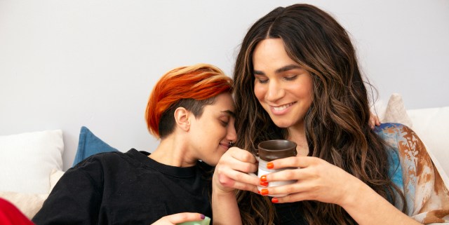 one person with short red, orange and yellow hair rests their nose on the shoulder of another person with longer brown hair, who is smiling with their eyes closed and holding a mug with both hands