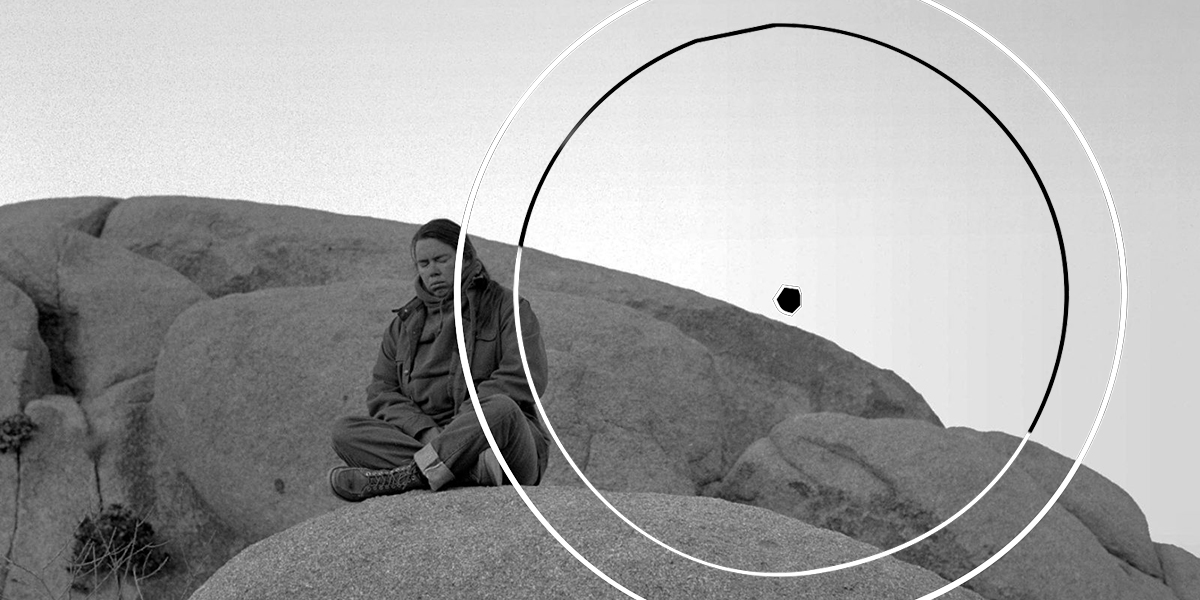 Pauline Oliveros is seated with eyes closed in a meditative pose on an outcropping of large, smooth rocks in a grainy black & white photo that looks like it was taken decades ago, when Oliveros was a young woman. The symbol of a perfect circle with a small dot in the center is superimposed over the photo.