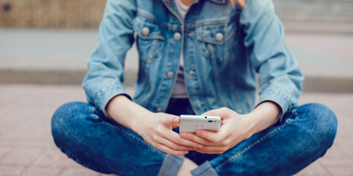 a seated person in dark wash denim jeans cuffed at the ankle and a denim jacket scrolls through their phone