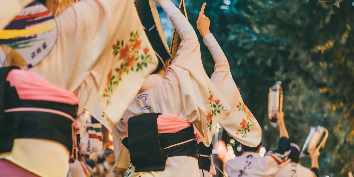 from the back, a line of three women in kimonos perform a Buddhist obon festival dance. the sleeves of their kimonos are decorated with flowers and their hands point to the sky. the dark blue and pink obi are tied on their backs.