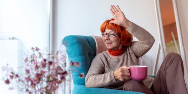 older woman sitting in teal armchair holding a pink mug and waving