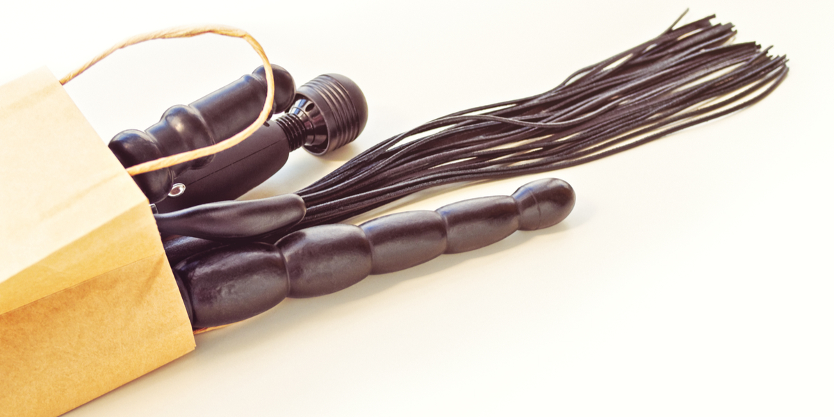 A range of black matte sex toys, from a wand vibrator to a flogger to a long penetrative wand, are seen poking out of a brown paper shopping bag.