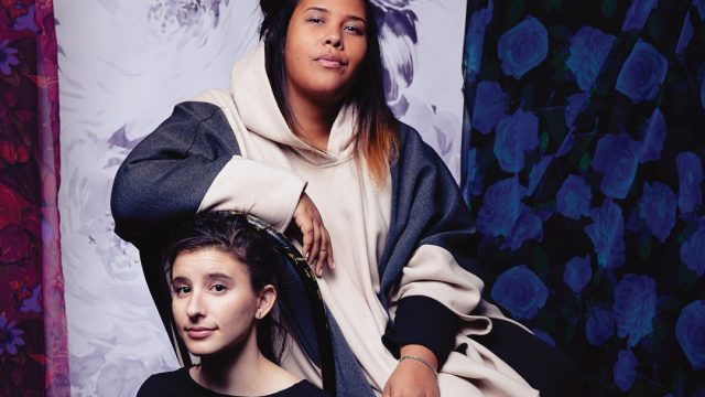 Emily Schorr Lesnick and Jamila Humphrie sit together against a multi-colored blue and purple background. Emily, who is white with curly brunette hair, sits on a chair while Jamila, who is black, rests in a higher chair above the top of her head.