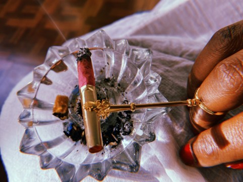 photo of a hand, fingernails painted red. A ring cigar holder is on one finger and it is holding a pink and gold cigarette.