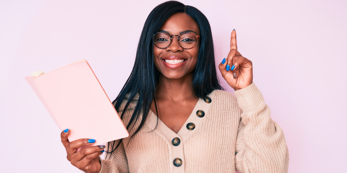A black woman stands against a pink background in a chunky knit sweater and holding up a pink book. Her face is proud and she has a single finger pointed to the air as if to say "A-ha! I have discovered something!"