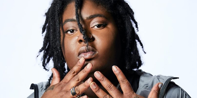 Chika is photographed against a plain white wall. Her locks fall casually into her face and she has her hands out in front of her.