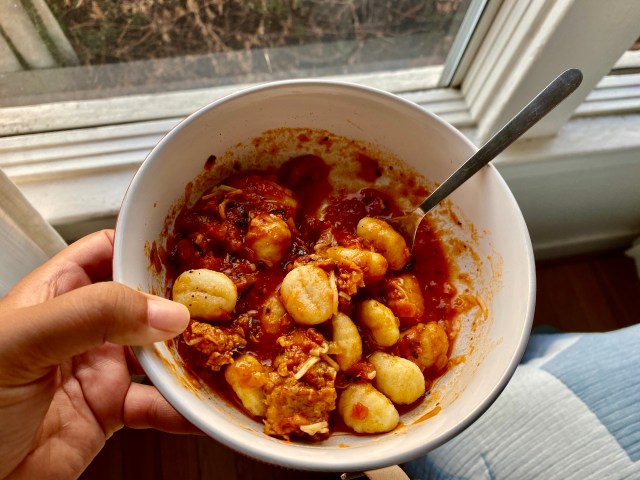 a brown hand holds a bowl of gnocchi mixed up in a red meat sauce