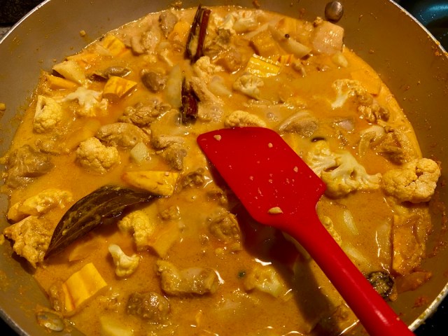 a pan with a yellow korma simmering around cauliflower, potatoes, chicken and squash