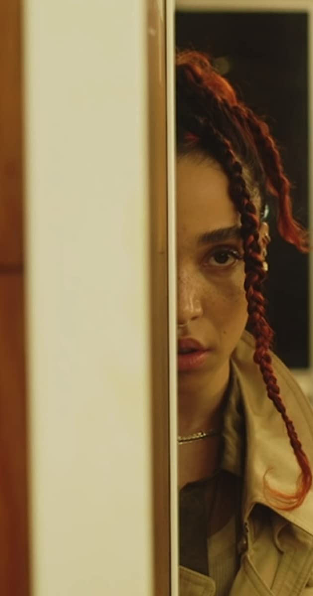 A still of FKA Twigs with braided red tinged hair standing behind a wall revealing only half of her face. The Still is from her music video "Sad Day"