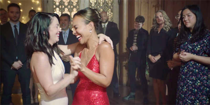 A still from the Netflix movie "A New York Christmas Wedding" where Jenny and her new-wife Gabrielle dance together at their reception. Jenny is an Afro-Latina in a sparkly red dress with her hair swept into a bun and Gabrielle is white, in a white pantsuit, and her dark hair down her back.