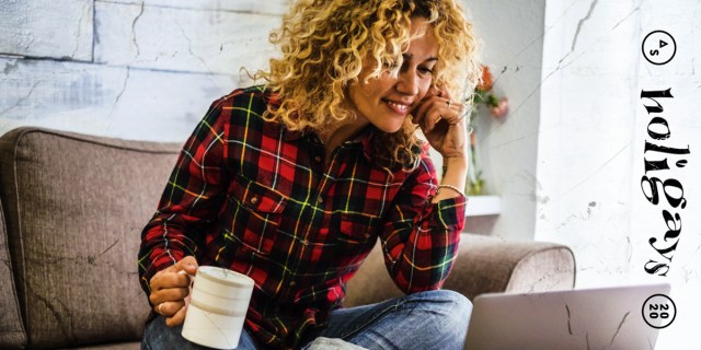 a smiling person in a flannel with curly hair, sits on the couch, holding a mug and leaning toward a laptop screen