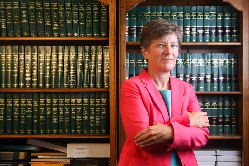 Mary Bonauto, Esq., Civil Rights Director at GLAD poses in front of a legal bookshelf in a pink blazer with her arms crossed.