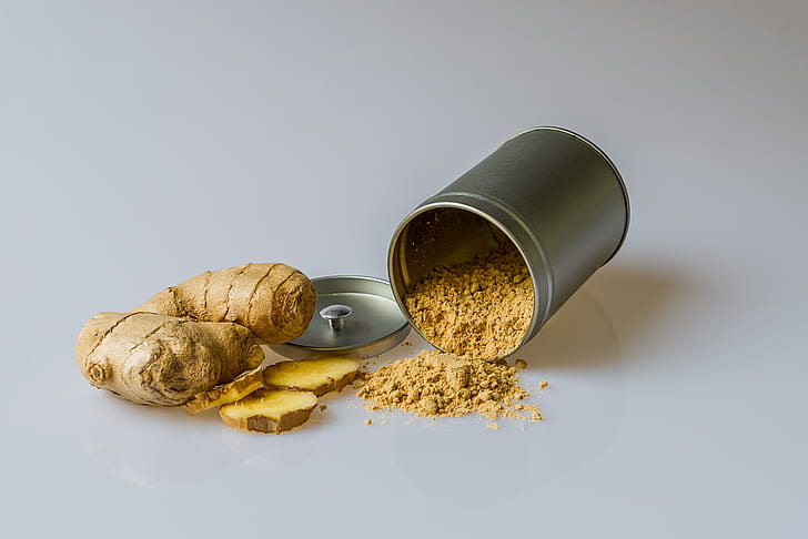 a whole ginger root with some pieces sliced off next to a fallen canister of ginger powder with its contents spilling out