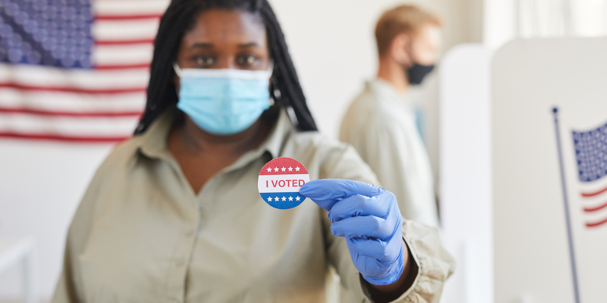 a black woman in a blue mask and gloves holds up a sticker that says "I voted"
