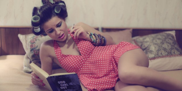 A woman wearing rollers and a red polka dot dress reclines on a bed with a cigarette, reading a copy of ODD GIRL OUT intently