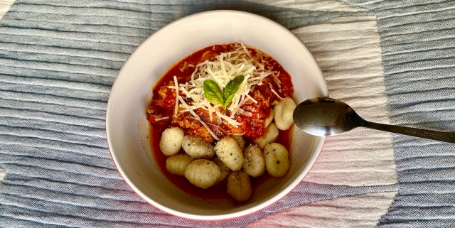 a bowl of gnocchi half covered in a red meat sauce topped with shredded cheese and a sprig of basil