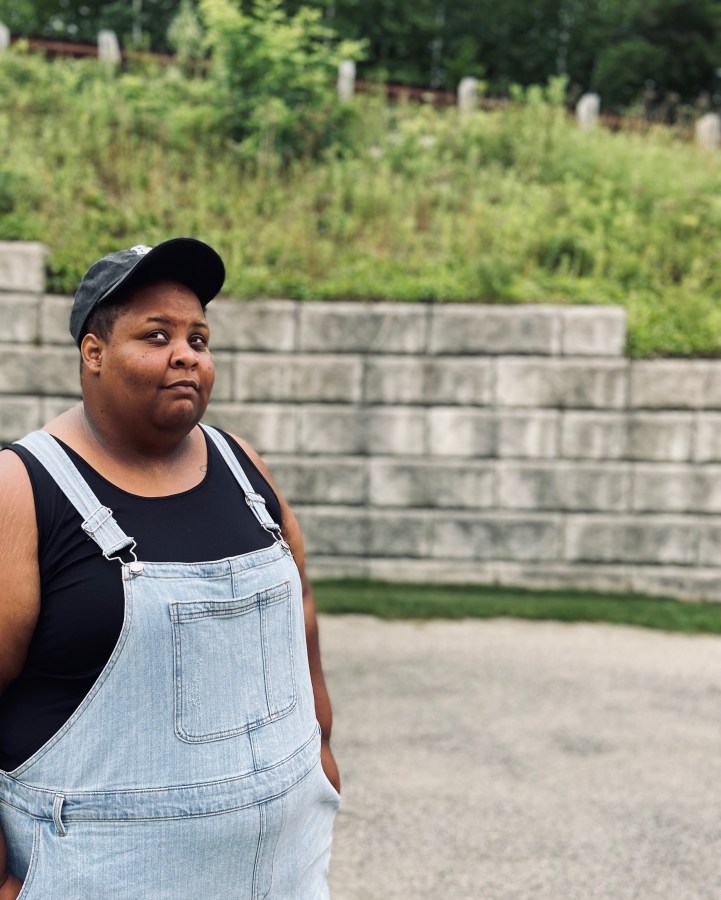 photo of a fat, black person in a black tank top, overalls and a cap, looking chill