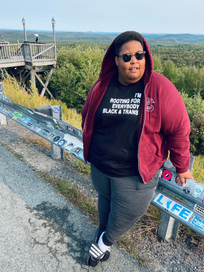 the author, a fat, Black person leans against the rail on the side of a mountain-side road beside a canyon. they're wearing a red hoodie with the hood up and sunglasses, and their shirt reads "I'm rooting for everyone Black & trans"