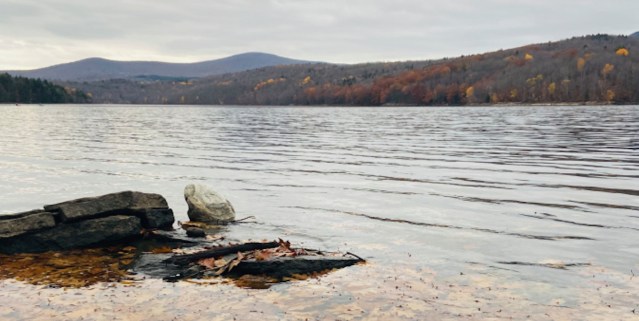 a photo of the placid top of a lake. in the foreground are few rocks emerge from the water and in the background are mountains with studded with colorful trees